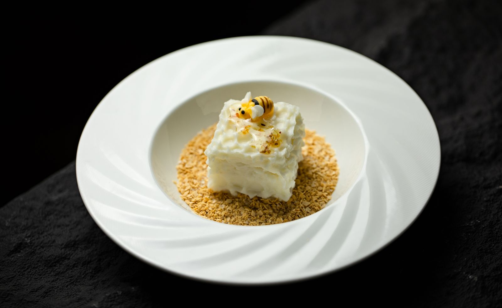 A gourmet white cake dessert topped with a cute edible bee on a bed of crispy oats.